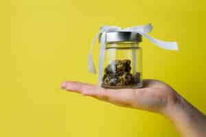 Female hand holding marijuana bud in glass jar with white ribbon on yellow background, close up. Concept of herbal and alternative medicine