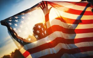 Closeup rear view of a couple holding American flag against summer sunset. They are visible as silhouettes.