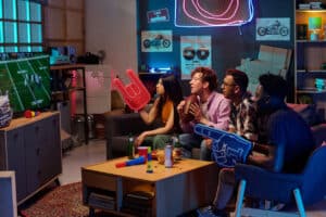 Group of young adult multiethnic friends sitting on couch watching football game on television, holding foam fingers and football, cheering and reacting to action on screen
